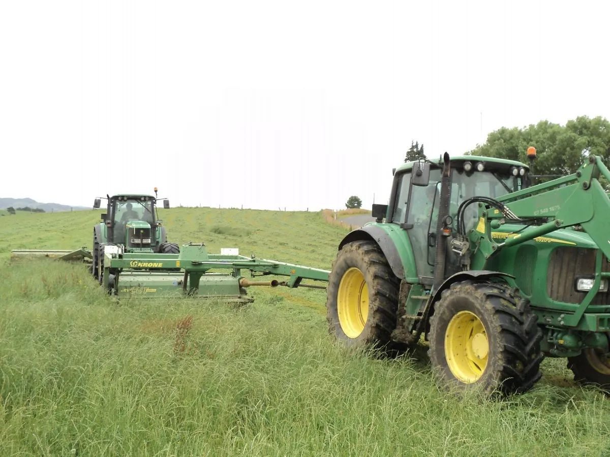 Two John Deere tractors mowing long grass, the far one pulling a wide Krone mower and the near one fitted with a front loader