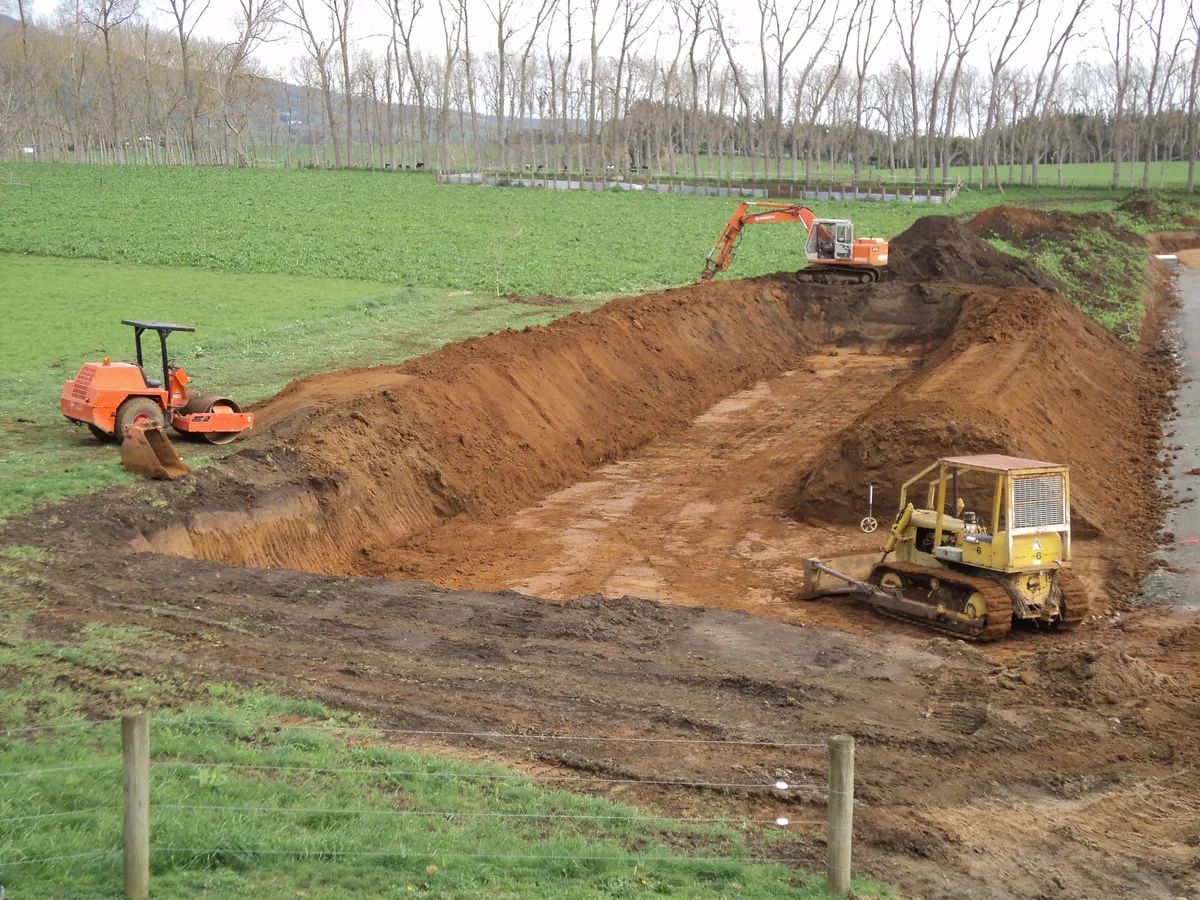 Bulldozer and excavator digging a large rectangular pit in a green paddock, with a roller parked on the bank and a row of poplars behind