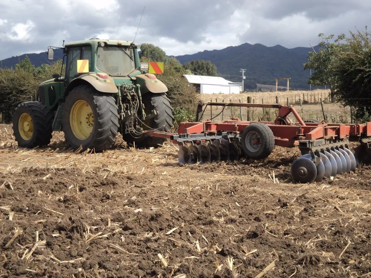 John Deere tractor pulling a set of red discs across a worked-up paddock of maize stubble, hills behind