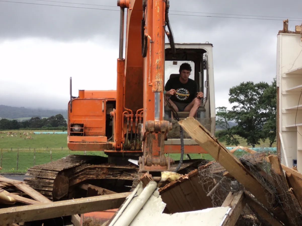Operator in the open cab of an orange excavator surrounded by broken timber from a building being pulled down