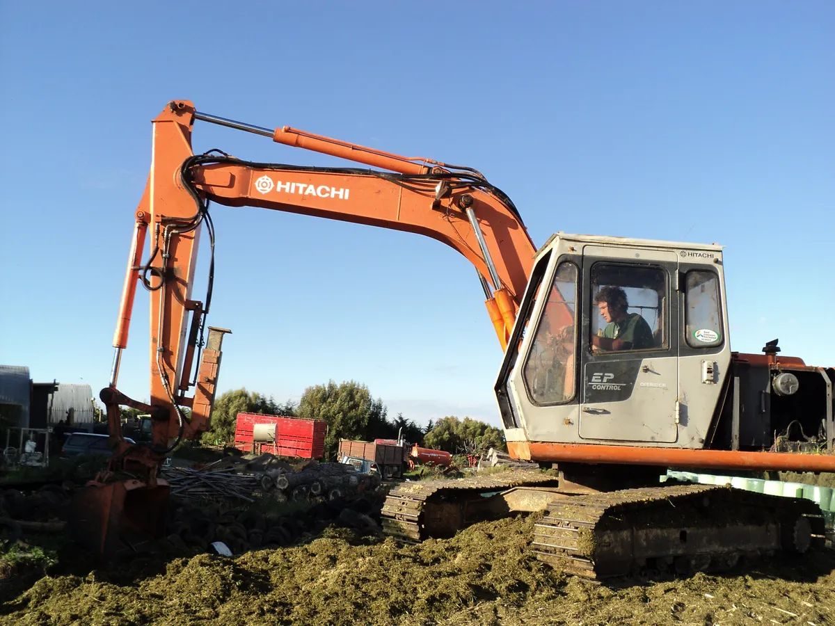 Orange Hitachi excavator with its operator in the cab, working on rough ground beside farm sheds