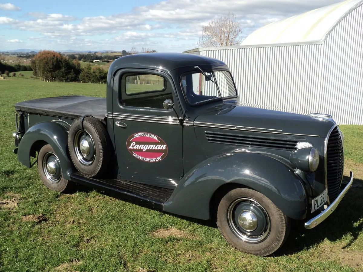 Restored dark green vintage Ford pickup with the Langman Agricultural Contracting oval on its door, parked on grass by a shed