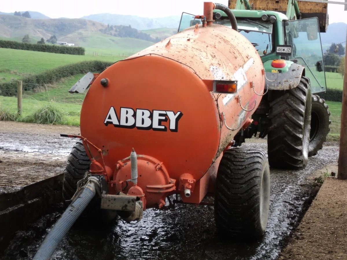 Orange Abbey effluent tanker behind a John Deere tractor, filling through a hose beside a concrete yard